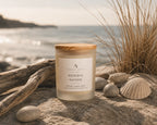 A jar candle labelled 'Nordic Shore' by Ardent North sits on a sandy beach with driftwood, seashells, and tall beach grass in the background.