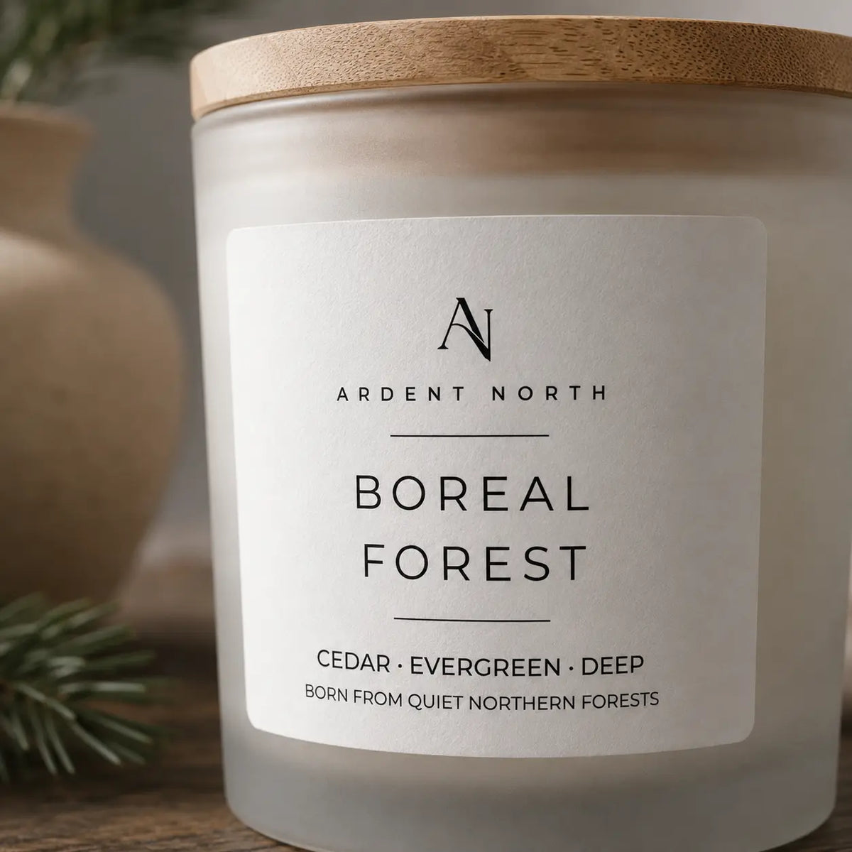 A close-up of an Ardent North candle jar with a light wood lid and a white label reading 'Boreal Forest' and 'Cedar • Evergreen • Deep'. The candle is photographed on a wooden surface with soft blurred background elements.