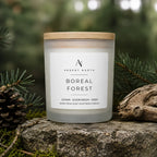 A frosted glass candle jar with a light wood lid sits on a stone outdoors, surrounded by pine branches, moss, and driftwood. The front label reads Ardent North Boreal Forest.