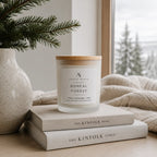 A frosted glass candle jar with a light wood lid sits on stacked neutral books on a wooden table, with a knit blanket, a vase with pine branches, and a softly blurred window view in the background. The label reads Ardent North and Boreal Forest.