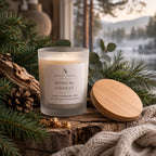 A frosted glass candle jar labelled 'Ardent North Boreal Forest' sits on a rustic wood surface with its round bamboo lid beside it, surrounded by pine branches, a pinecone, eucalyptus leaves, and a knitted blanket. A blurred outdoor winter forest scene appears in the background.