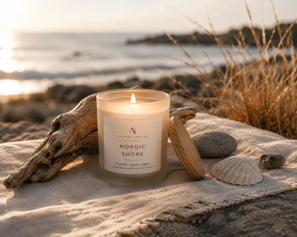 A lit glass jar candle labeled 'Nordic Shore' by Aesthetic House sits on a sandy beach with driftwood, seashells, and dry grass in the background.