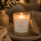 A lit jar candle labelled Nordic Shore, sits on a stone tray with its wooden lid beside it, in a softly styled home setting with warm lighting and a vase in the background.