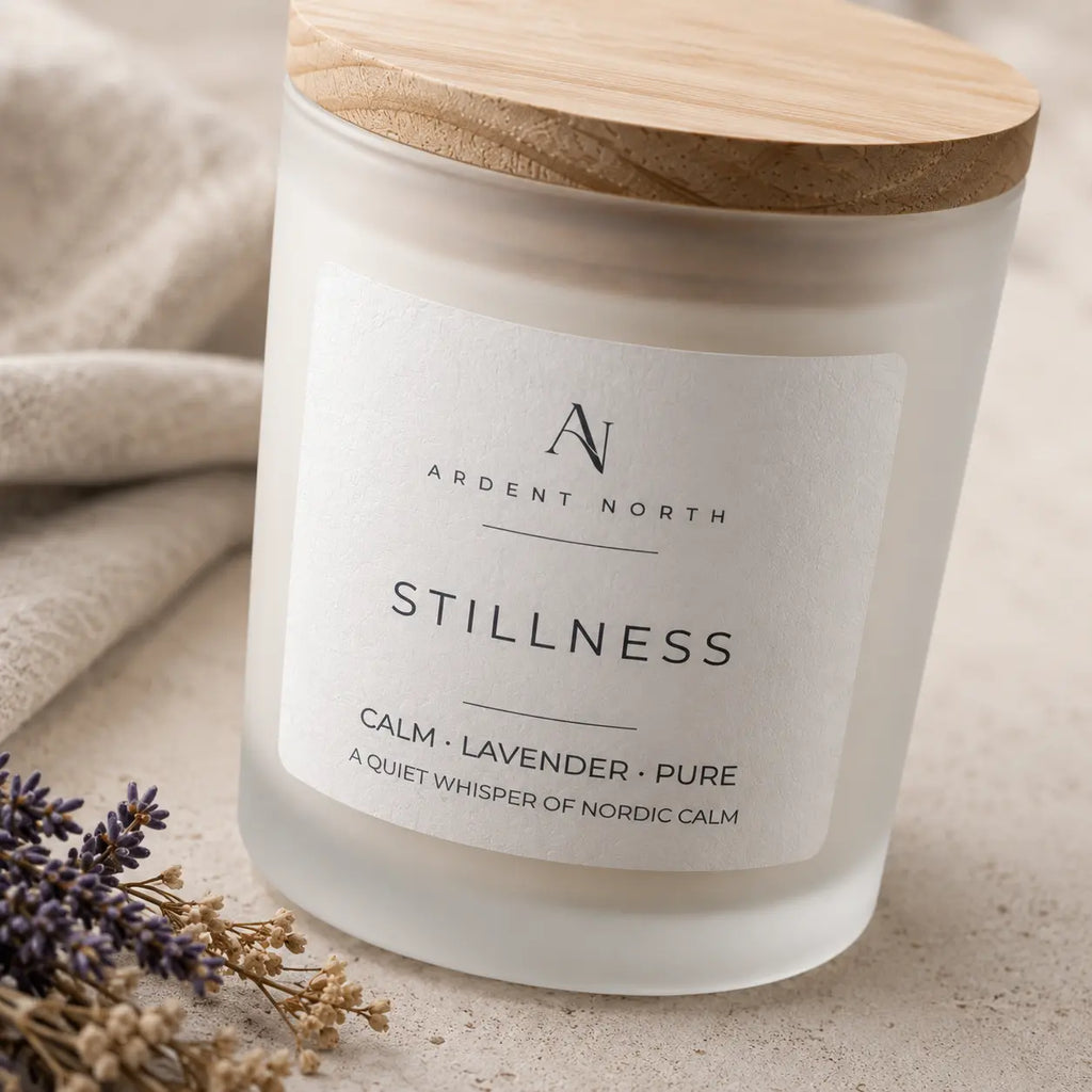 A frosted glass jar candle with a light wood lid and a white label reading 'Ardent North Stillness Calm • Lavender • Pure'. The candle is styled on a neutral textured surface with a beige cloth and dried lavender sprigs.