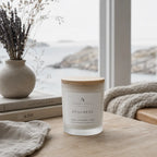 A frosted glass jar candle with a light wood lid sits on a wooden table in a softly styled room by a window. The label reads 'Ardent North' and 'Stillness Calm, Lavender, Pure'.