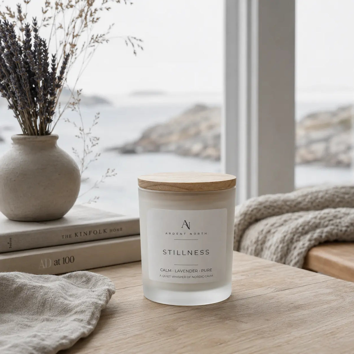 A frosted glass jar candle with a light wood lid sits on a wooden table in a softly styled room by a window. The label reads 'Ardent North' and 'Stillness Calm, Lavender, Pure'.