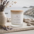 A frosted glass candle jar with a light wood lid and a white label reading 'Ardent North Stillness Calm - Lavender - Pure', styled on a stone surface with dried lavender, a ceramic vase, and a soft neutral fabric background with a blurred coastal scene.