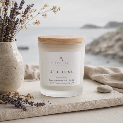 A frosted glass candle jar with a light wood lid and a white label reading 'Ardent North Stillness Calm - Lavender - Pure', styled on a stone surface with dried lavender, a ceramic vase, and a soft neutral fabric background with a blurred coastal scene.