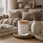 A frosted glass candle jar with a light wood lid sits on a wooden tray in a cosy living room setting. The label reads 'ARDENT NORTH' and 'STILLNESS', with scent details 'CALM, LAVENDER, PURE'.