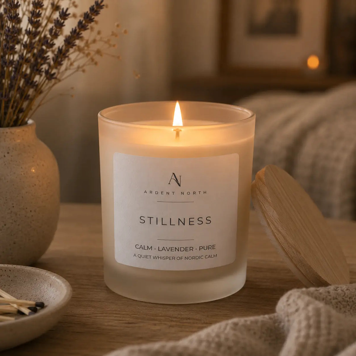 A lit candle in a frosted glass jar sits on a wooden surface in a cosy indoor setting. The label reads 'ARDENT NORTH' and 'STILLNESS' with 'CALM + LAVENDER + PURE' beneath it.