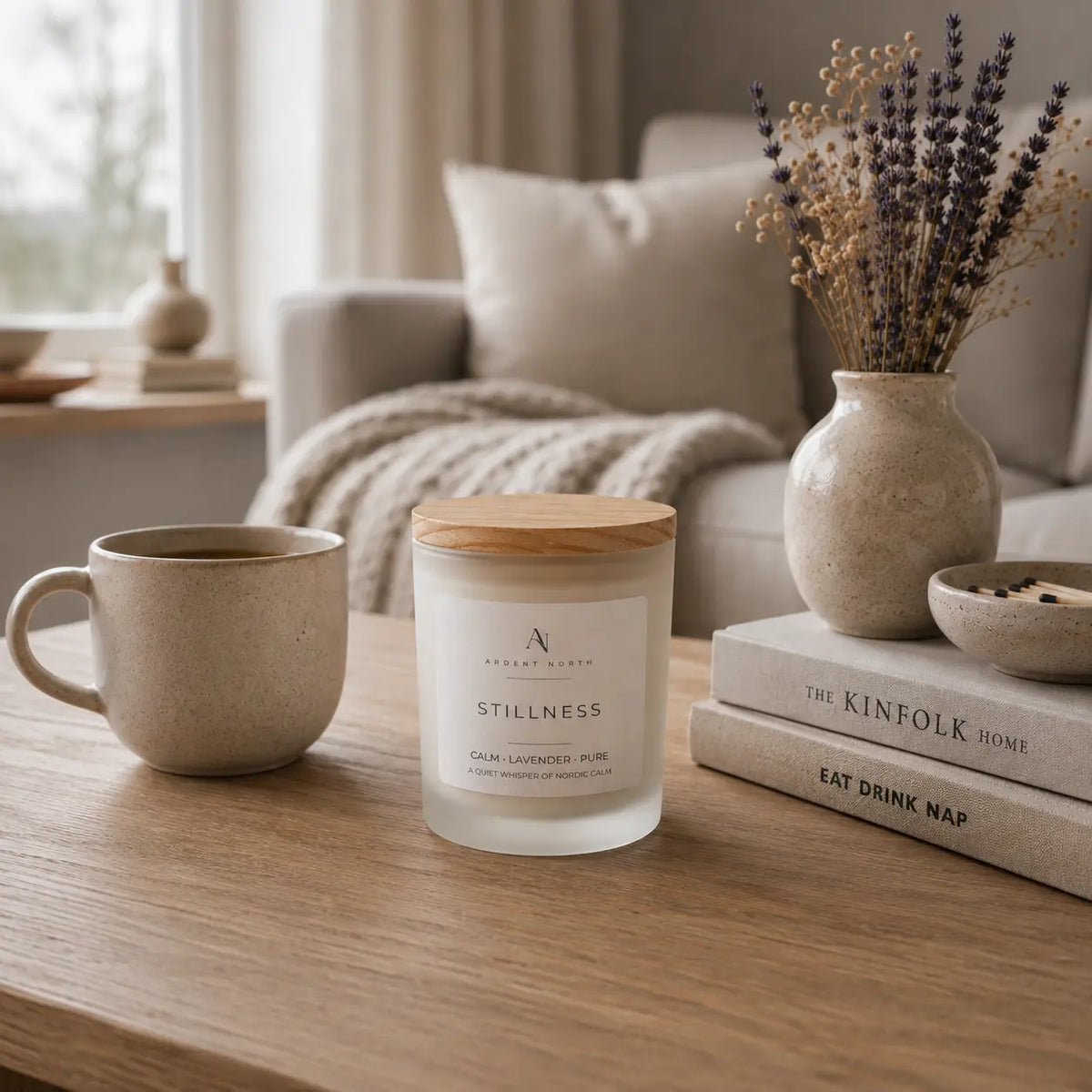 A frosted glass jar candle with a light wood lid sits on a wooden table in a cosy living room setting, styled with a ceramic mug, books, and a vase of dried lavender.